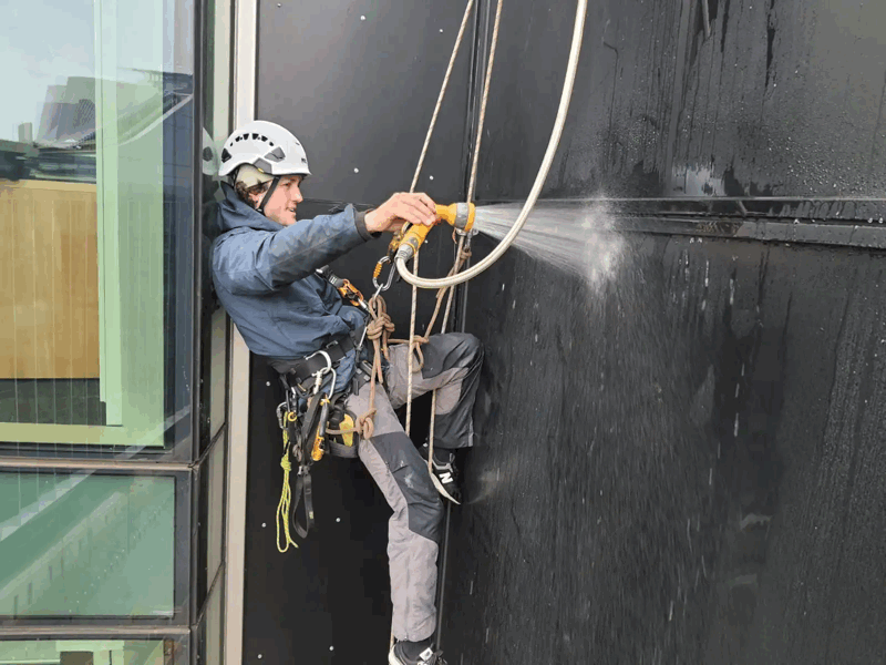 Rope access technician in harness performs a hose test on dark facade cladding panels
