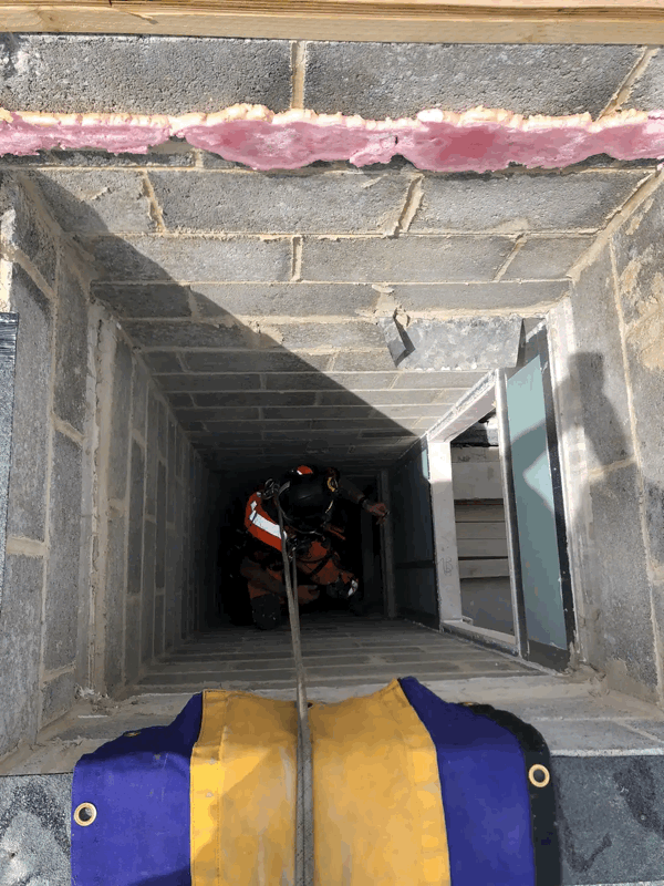 Technician in orange protective suit inspecting a narrow masonry cavity behind exterior cladding wall