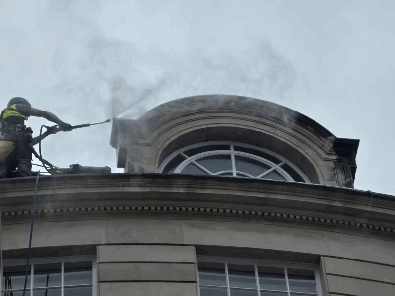 Worker on rooftop uses pressure washer to clean steam off a stone dormer window on a historic building facade