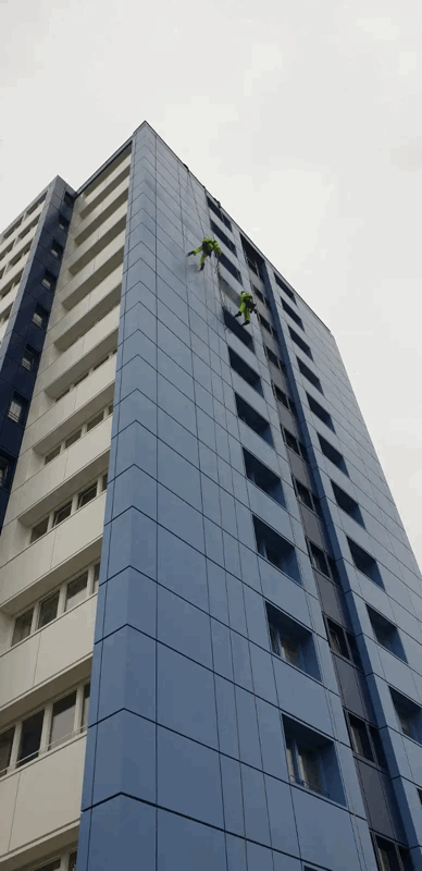 Two rope access technicians inspect blue metal cladding panels on a tall high-rise building facade.