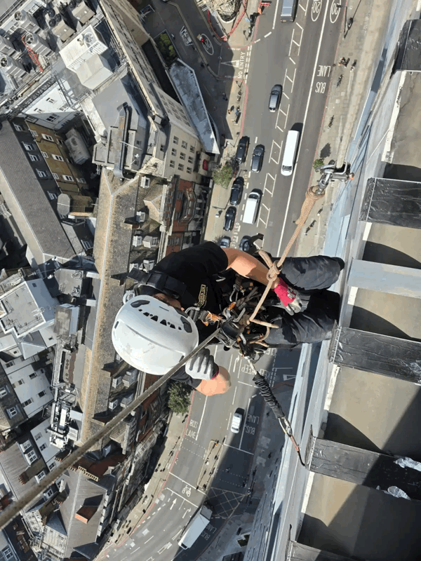 Rope access technician in helmet and harness abseils down a building facade high above a city street