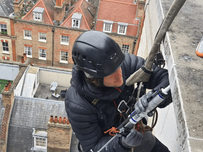 Rope access technician in helmet and harness applies sealant to a building facade high above a rooftop