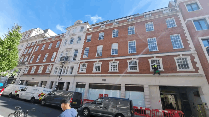 A technician on rope access equipment inspects the brick facade of a multi-storey London building above a street with parked