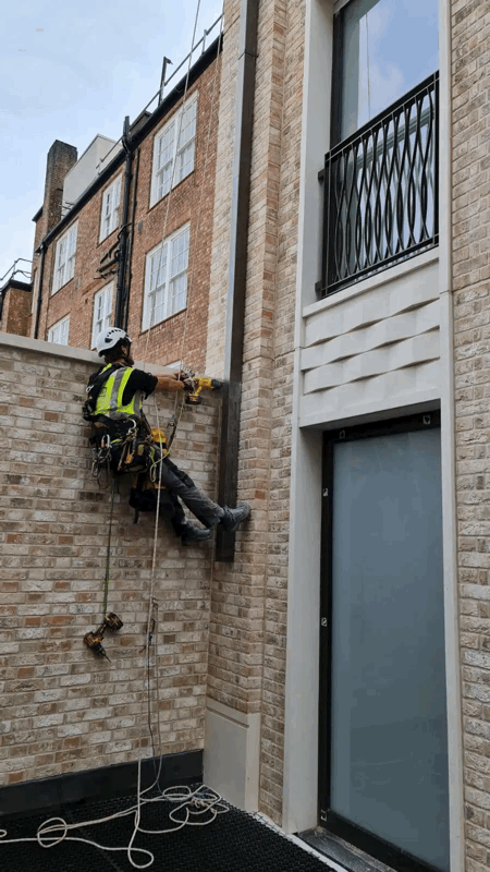 Rope access technician in harness inspects brick slip cladding on a building facade using hand tools.