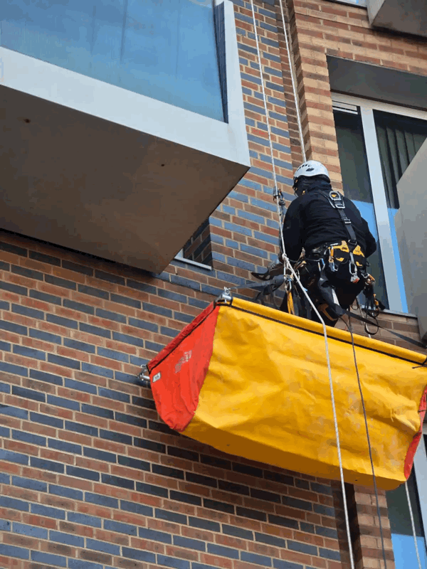 Rope access technician in harness inspects brick cladding facade beside a yellow protective debris chute