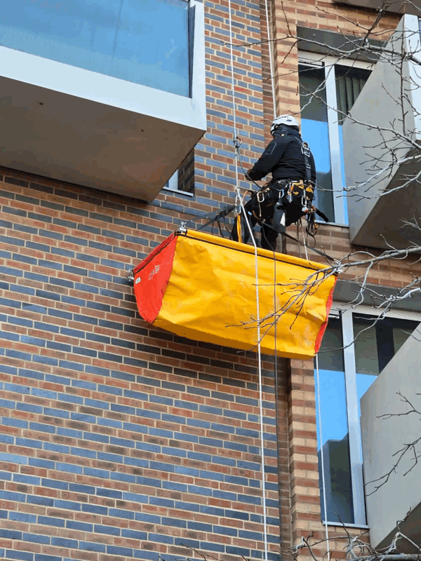 Rope Access Cladding Inspection on Brick Facade Rope access technician abseils down a brick clad building beside a yellow debris chute during cladding inspection