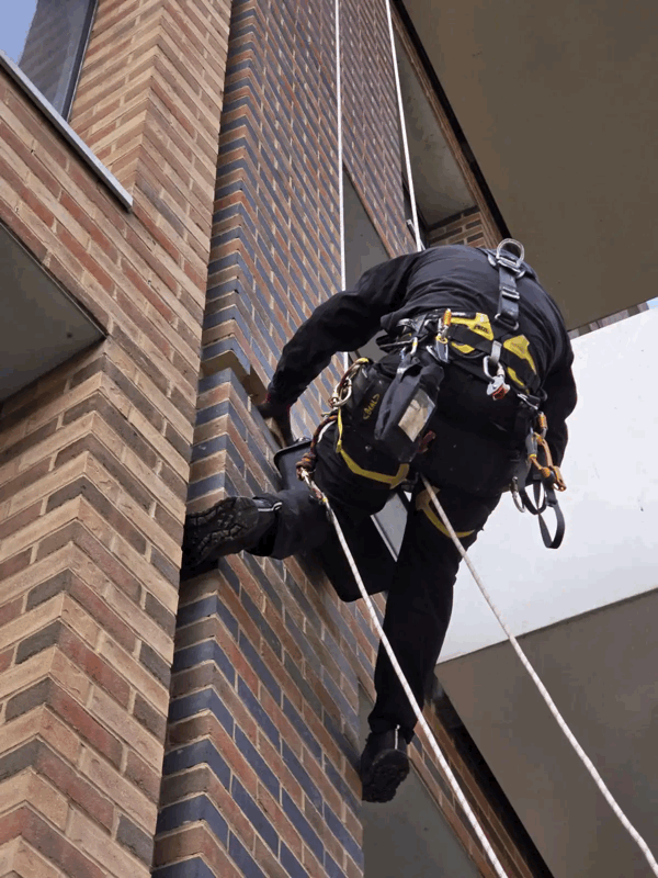 Rope access technician in black harness inspects brick slip cladding on a high-rise building facade
