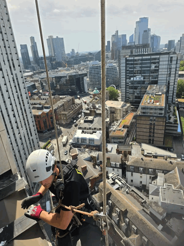 Rope access technician in helmet and harness abseils down a high-rise building during a cladding inspection above a city