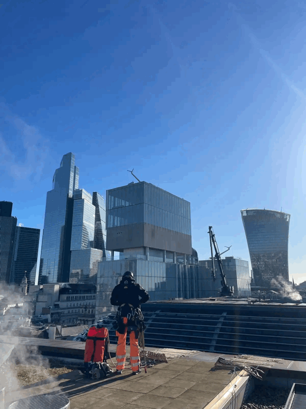 Rope access technician in orange safety trousers inspects rooftop cladding against London's city skyline.