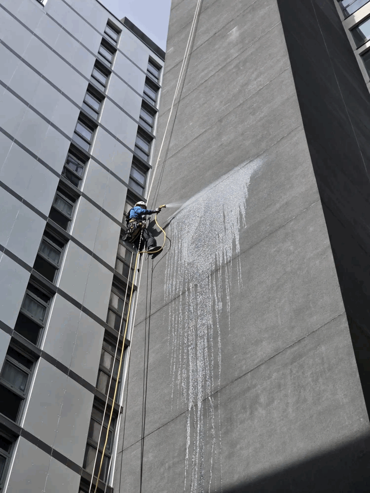 Rope access technician cleans grey cladding panels on a high-rise building facade with a pressure hose.