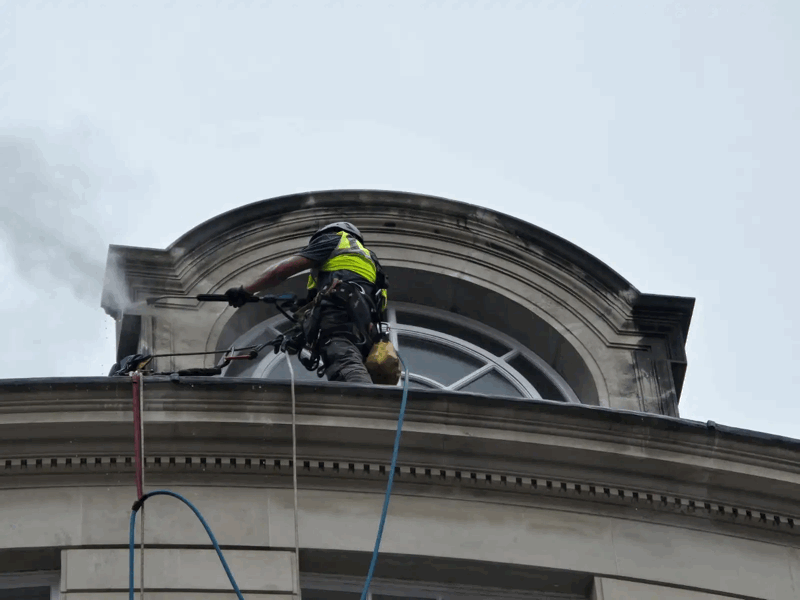 rope access cladding inspection — Rope Access Cleaning of Ornate Stone Rope access technician in high-visibility vest pressure washing a stone building facade below an arched window