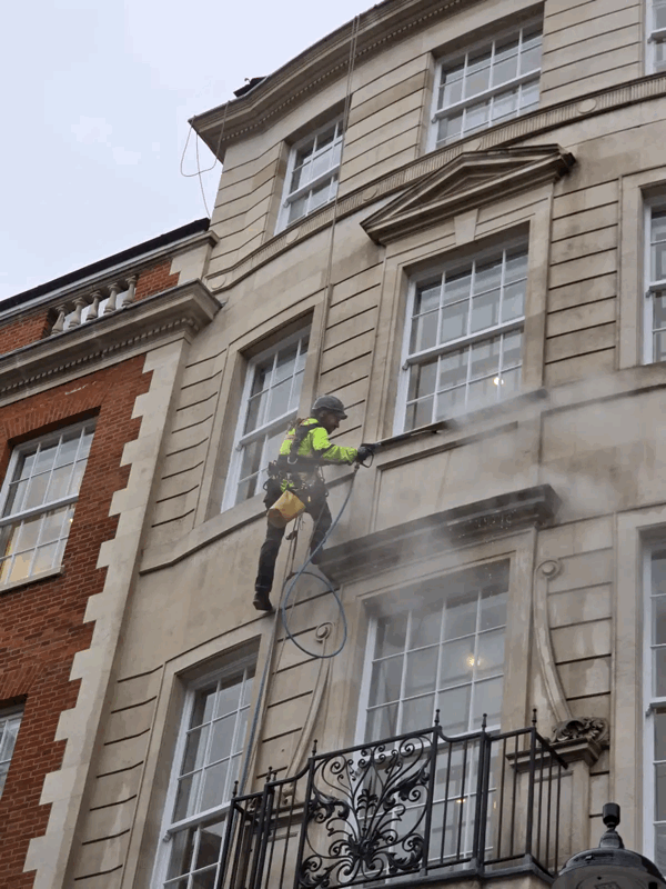 Rope access technician in high-vis gear cleans upper-storey windows on a stone building facade using a hose.