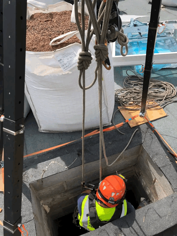 Rope access technician in orange helmet descends through a roof hatch opening during a building inspection