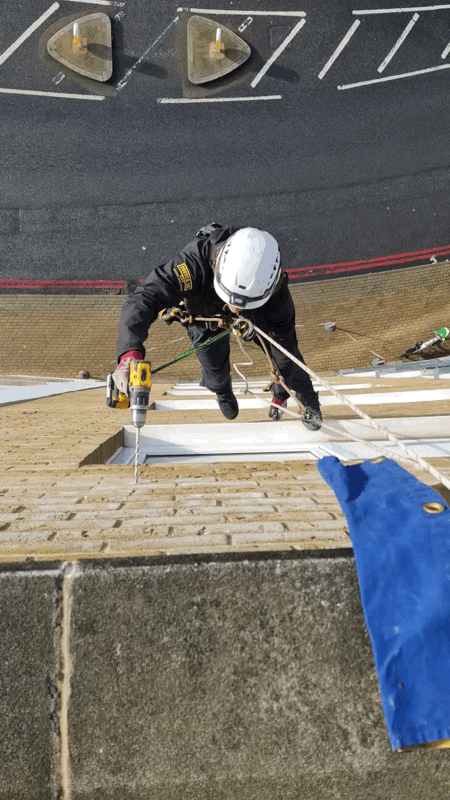 Rope access technician in helmet drills into beige brick-slip cladding panel on a building facade
