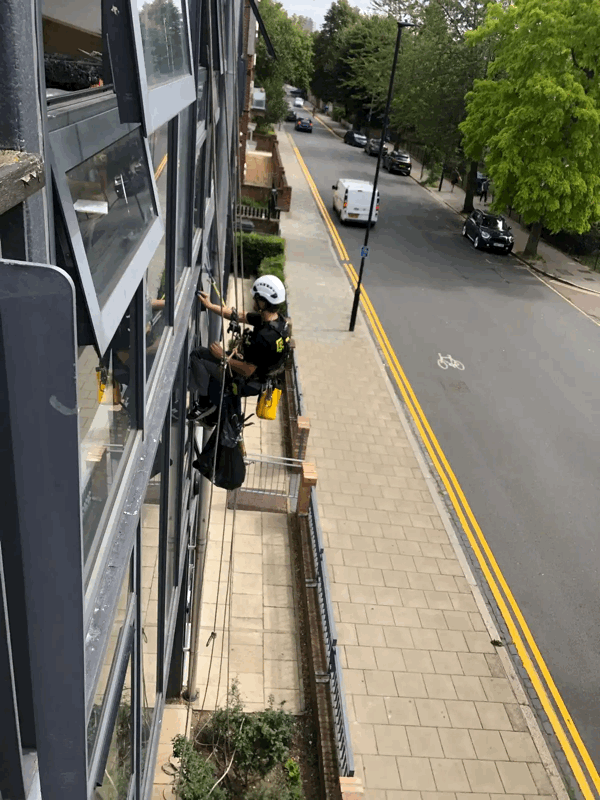 Rope access technician in helmet and harness inspects window cladding on a multi-storey building facade above a street