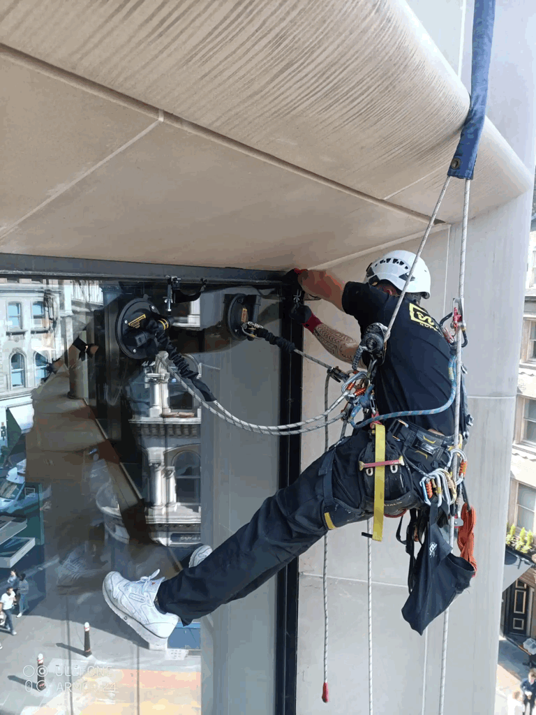 Rope access technician in harness inspects building facade cladding joint high above a city street