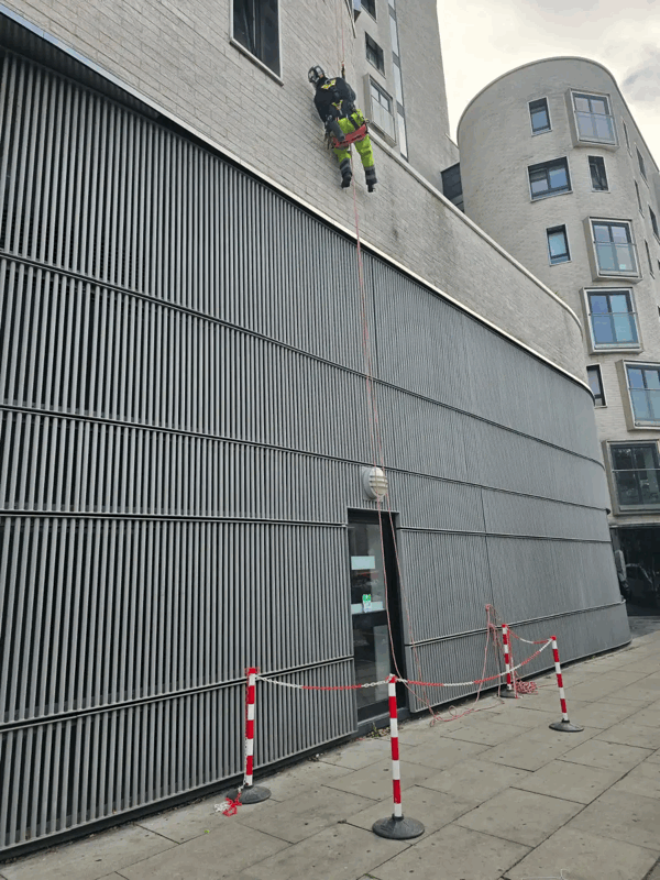 Rope access technician in high-vis gear inspects grey metal cladding on a building facade above a barriered walkway