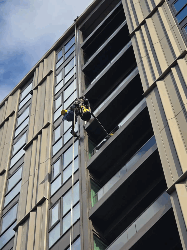 Rope access technician in harness inspecting beige panel cladding on a high-rise building facade