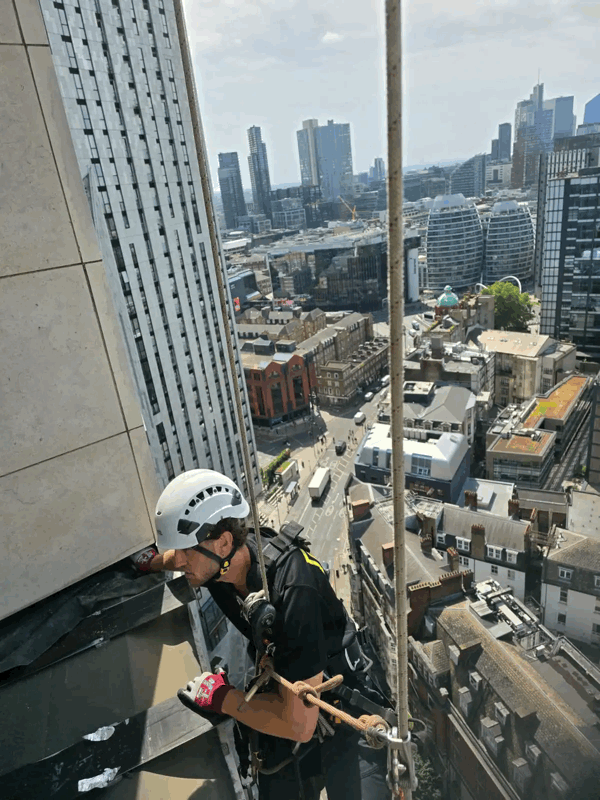 Rope access technician in helmet and harness inspecting stone cladding panels on a high-rise building above a city skyline