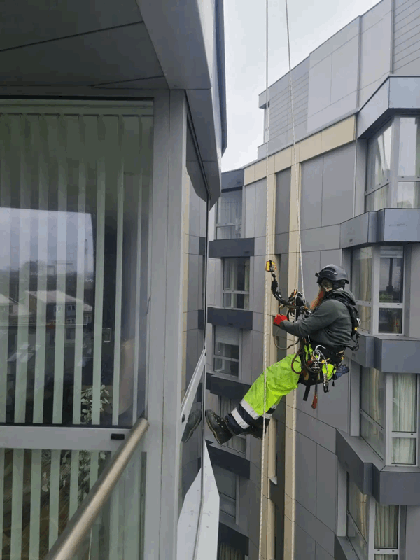 Rope access technician in harness inspects grey rainscreen cladding panels on a high-rise building facade