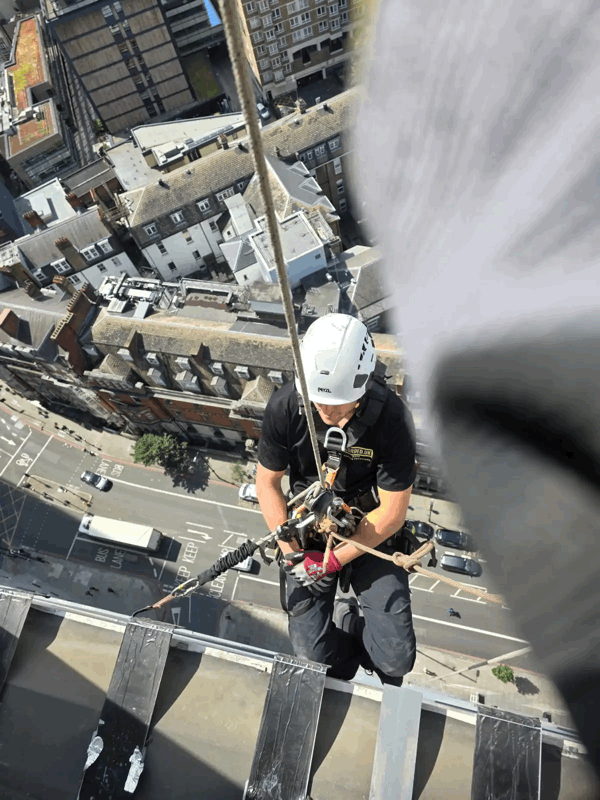 Rope access technician in helmet and harness inspecting building cladding high above a city street