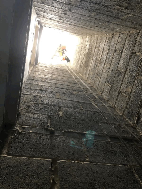 Rope access technician descending a narrow cladding cavity shaft with daylight visible at the top opening