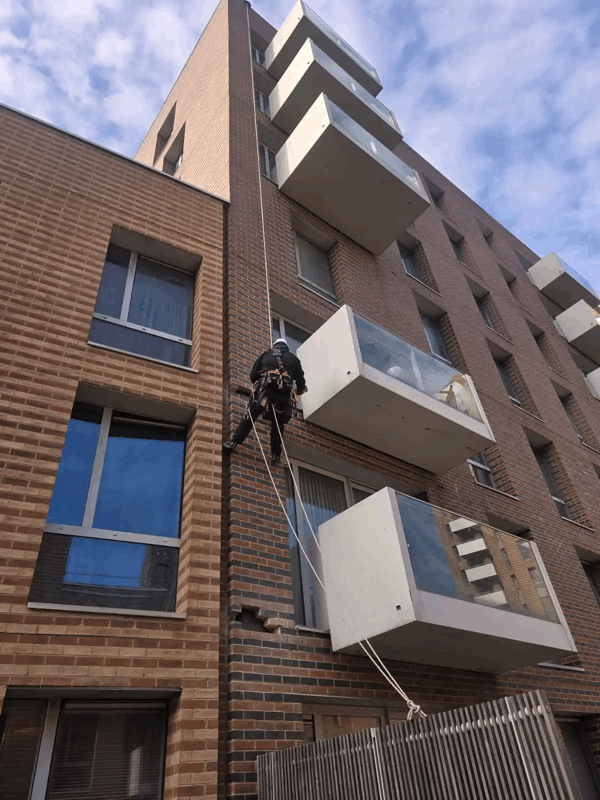 Rope access technician in harness inspecting brick facade and white balcony cladding on a high rise building