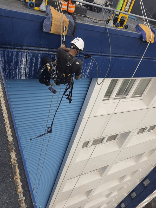 Rope access technician in harness inspecting blue metal cladding panels on a high-rise building facade