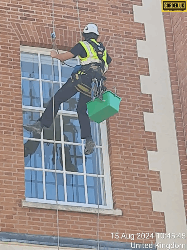 Rope access technician in harness and helmet inspecting a brick building facade with tools bag
