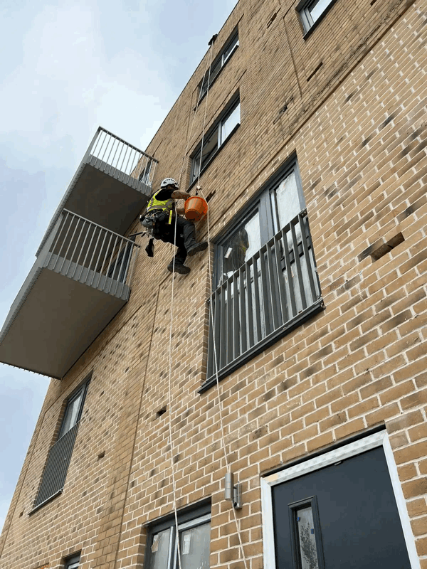 Rope access technician in harness inspects a brick-clad residential building facade near balconies.