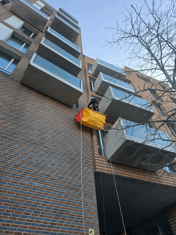 Rope access technician in harness inspects a brick-clad high-rise facade beside glass balconies.