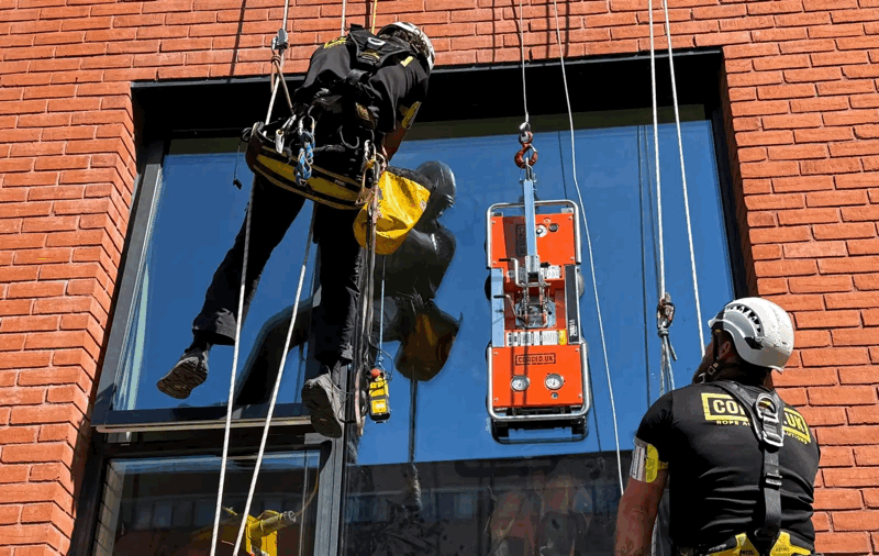 Rope access technician in harness inspects glazing on a red brick building while colleague monitors from below