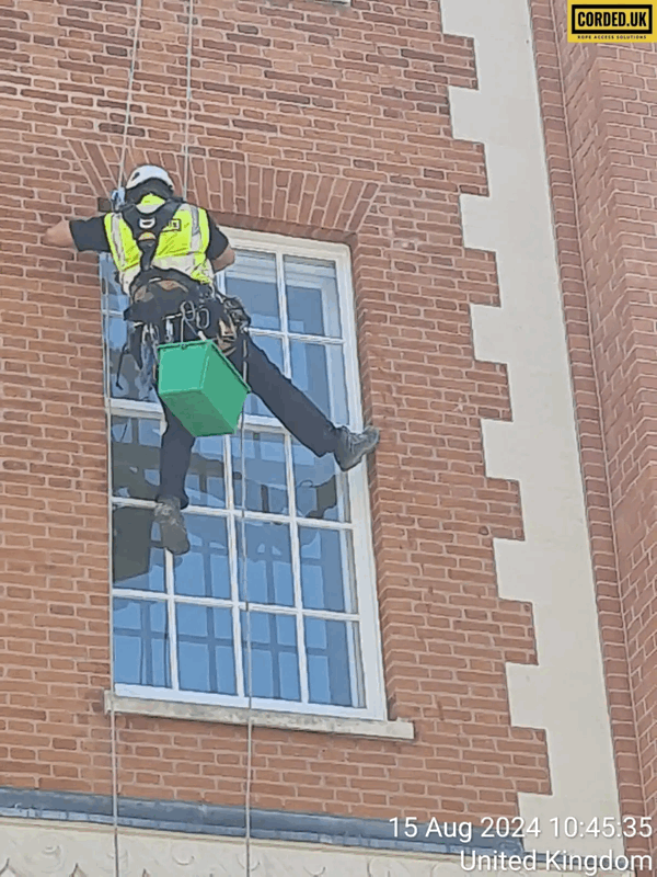 Rope access technician in high-vis harness inspects a brick building facade beside a window, carrying a green equipment box.