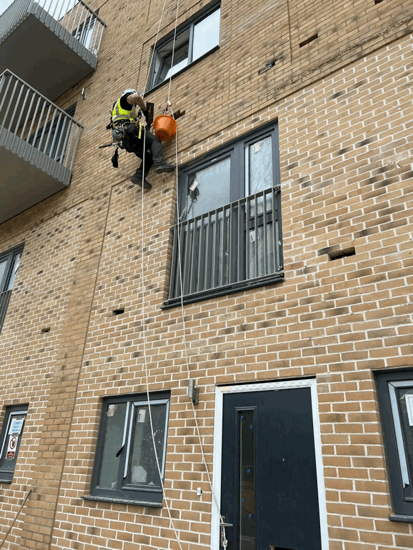 Rope access technician in harness abseiling down a brick apartment building during a facade inspection