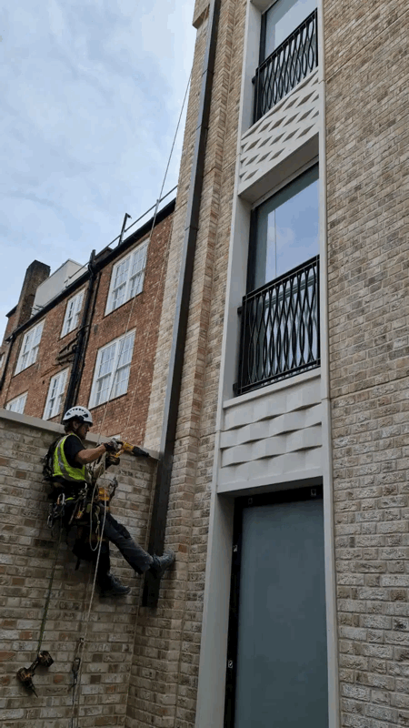 Rope access technician in helmet and harness inspects brick cladding on a multi-storey building facade.