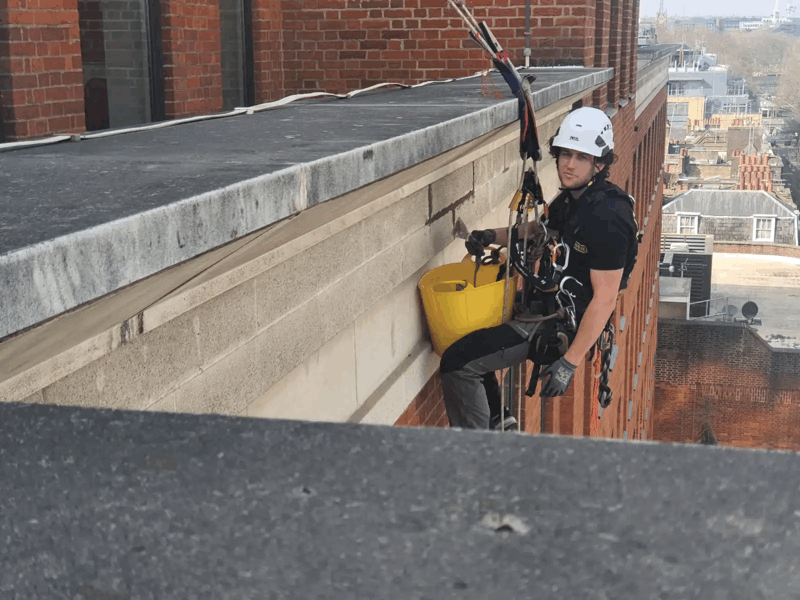 Rope access technician in helmet and harness inspecting a brick building facade high above the city.