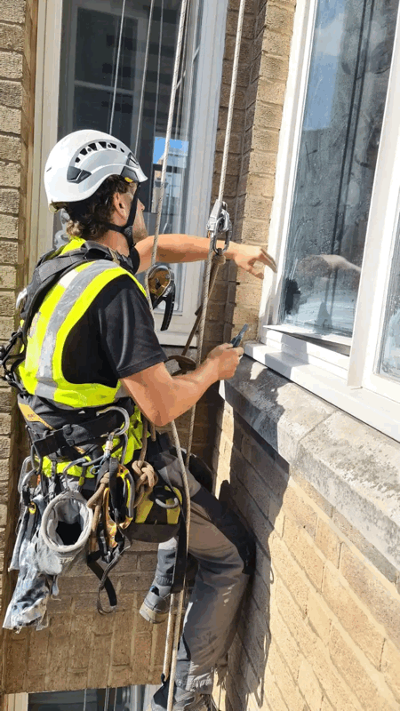 Rope access technician in harness and helmet inspects a window and brick cladding on a building facade