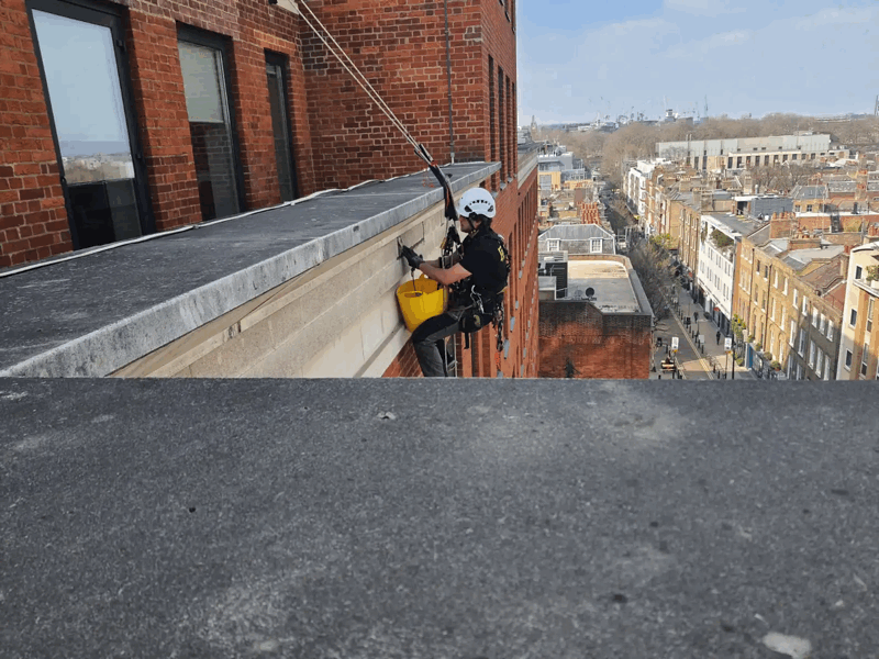 Rope access technician in helmet and harness inspects a brick building facade high above a London street.