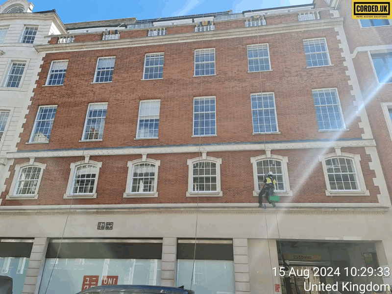 Rope access technician in harness inspecting upper-floor windows on a red brick building facade in London.