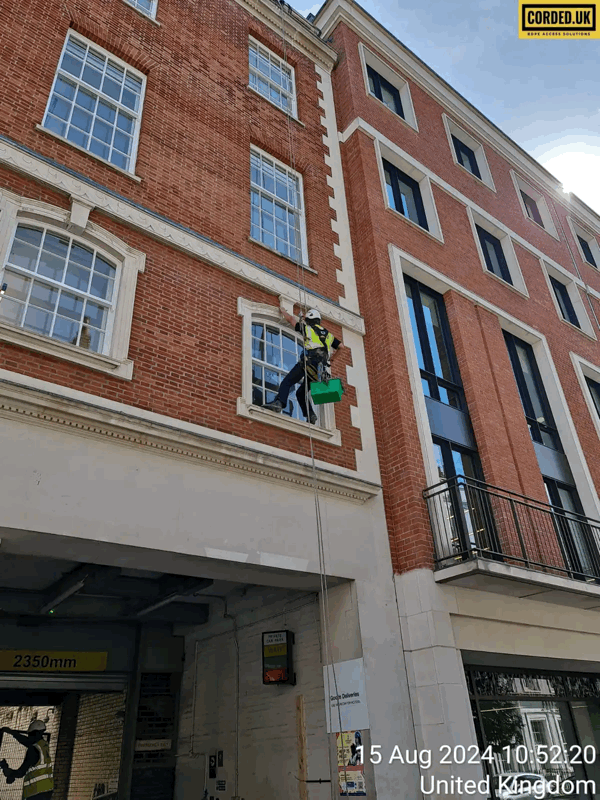 Rope access technician in harness inspects a brick building facade beside a window with a green tool box