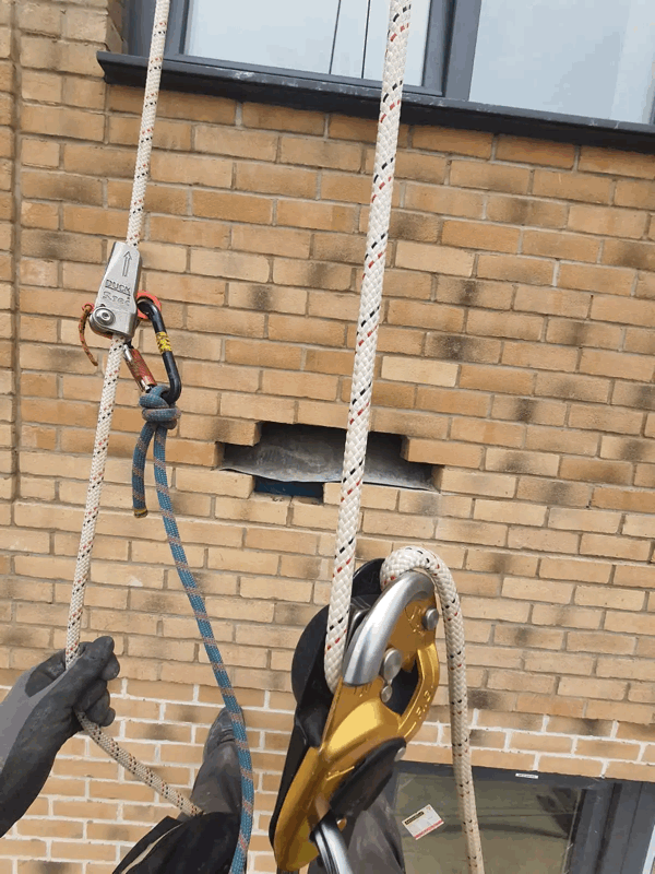 Rope access technician using ropes and descender gear to inspect a brick-clad building facade with an opened inspection panel