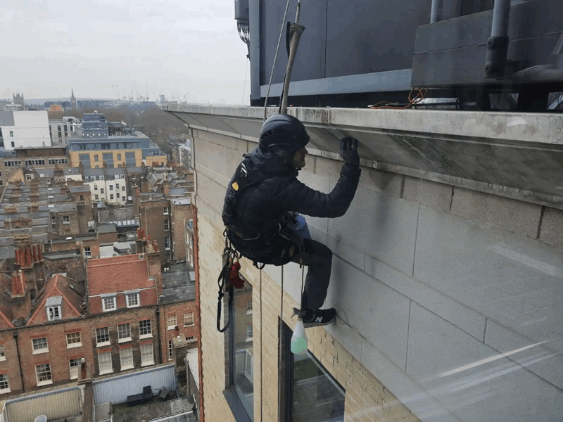 Rope access technician in harness inspecting stone cladding on a high-rise building facade above a city skyline