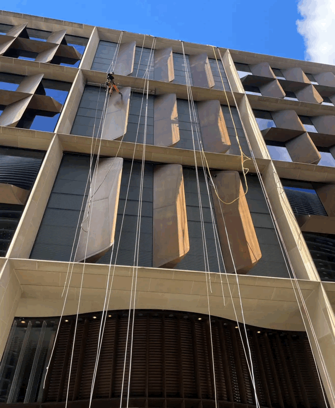 Rope access technician inspects tan cladding panels on a high-rise building facade under a blue sky
