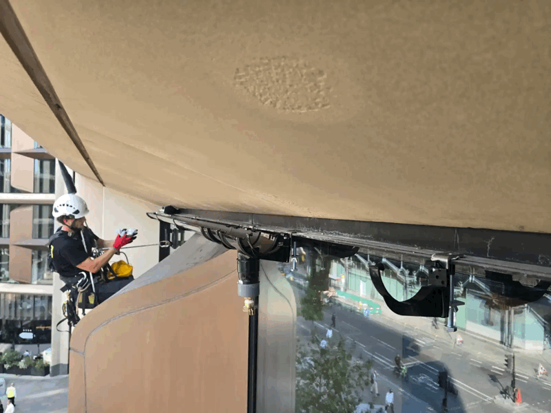 Rope access technician in helmet inspecting beige cladding soffit and fixings high on a building facade