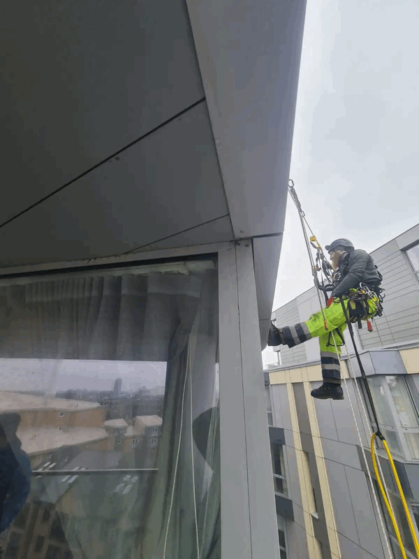 Rope access technician in harness inspects grey rainscreen cladding on a high-rise building facade