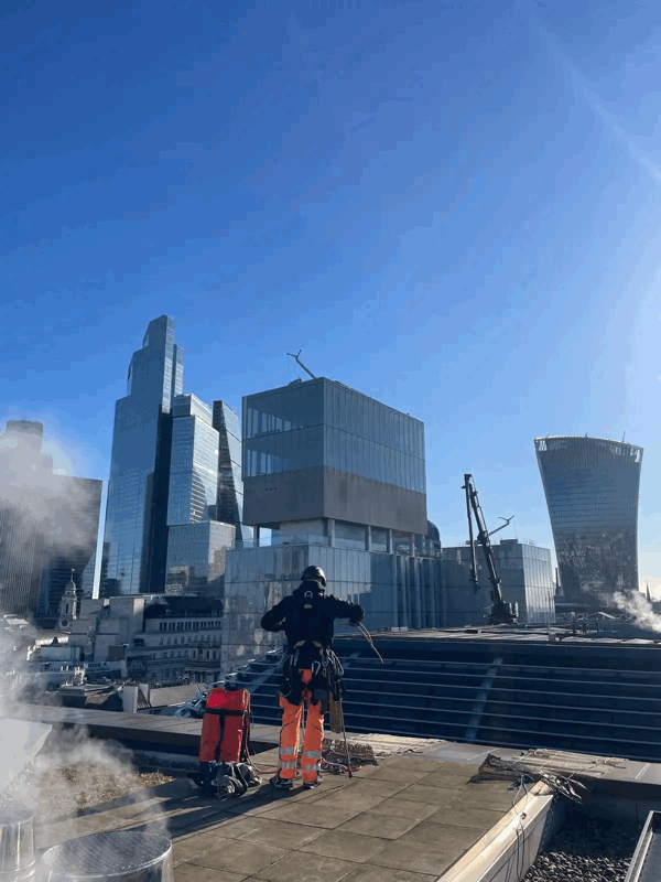 Rope access technician in harness inspects glazed building facade on a rooftop amid London skyscrapers.