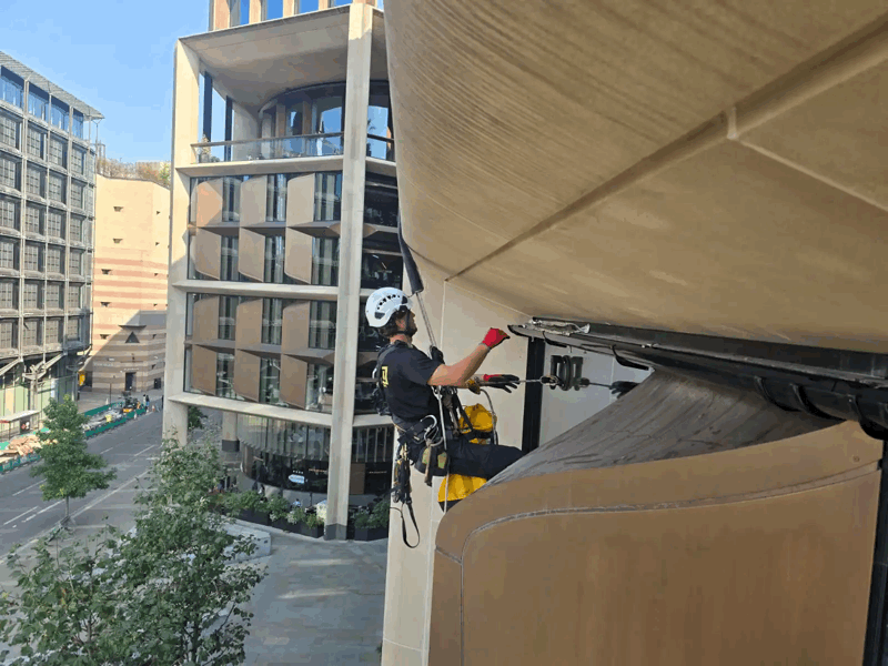 Rope access technician in helmet and harness inspects curved beige cladding panels on a high-rise building facade.