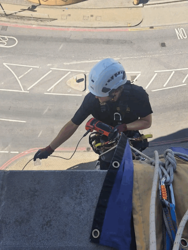 Rope access technician in helmet inspects grey cladding panel on building facade above street