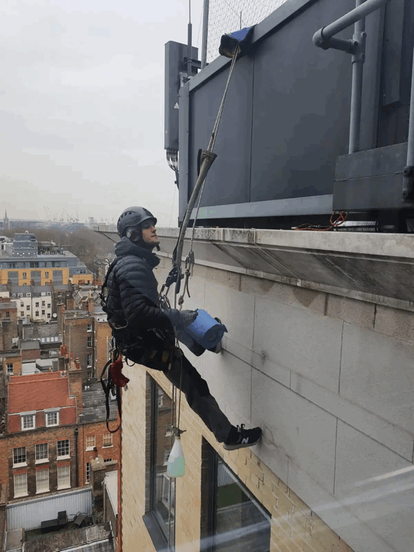 Rope access technician in helmet and harness inspects stone facade cladding on a high-rise building above a city street