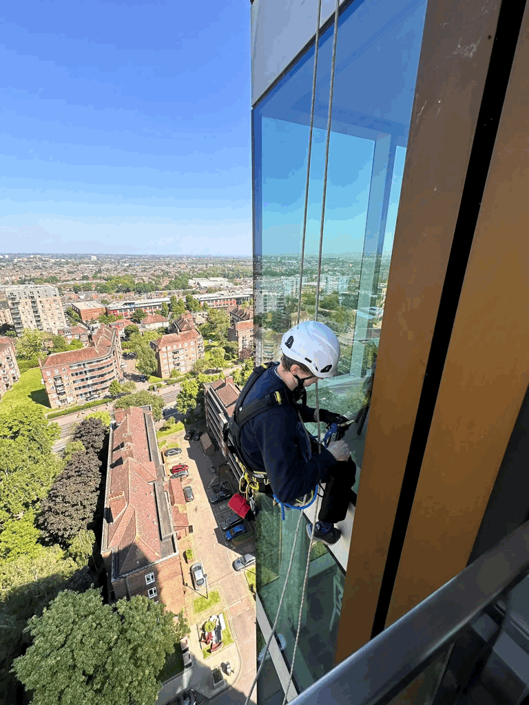 Rope access technician in helmet and harness inspects glass panel cladding on a high-rise building above a city skyline.